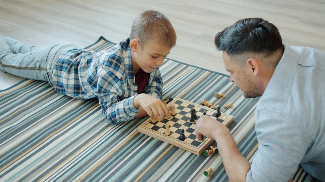 Smart child is playing chess board game with father middle-aged man in casual clothing spending free time at home. Leisure, family and childhood concept.